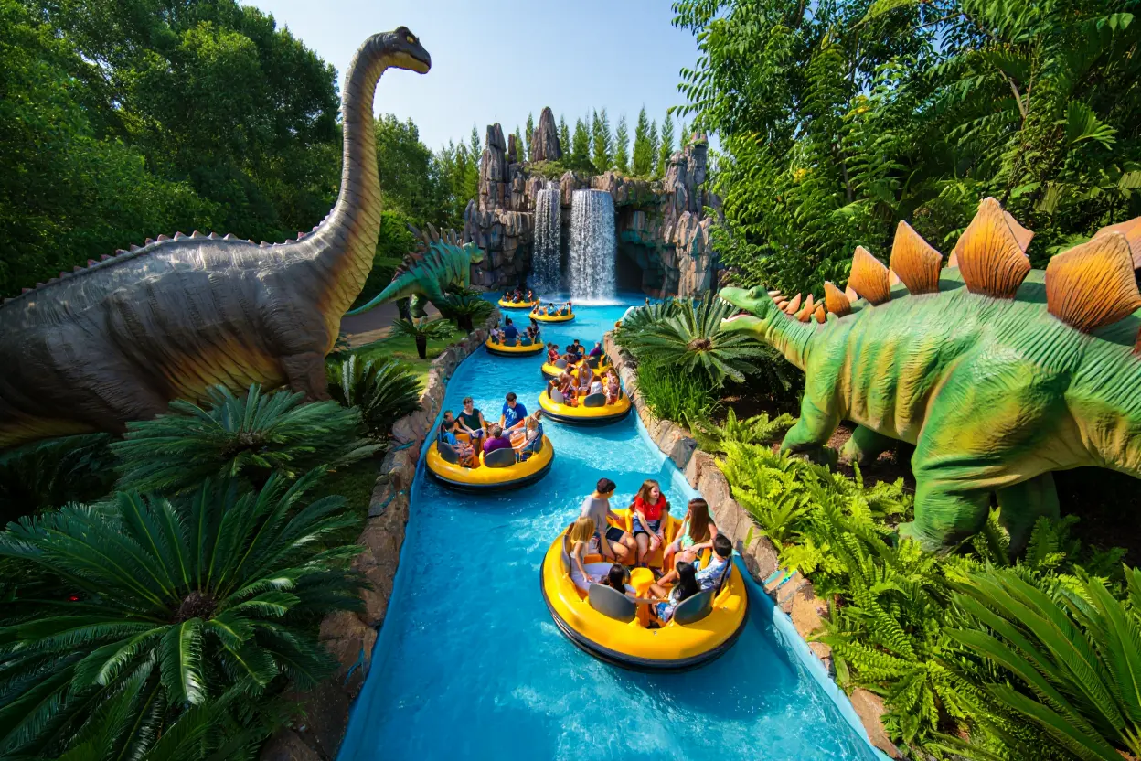 Des visiteurs à bord de bouées circulaires jaunes naviguent dans une rivière au milieu de dinosaures animés et de végétation tropicale luxuriante.